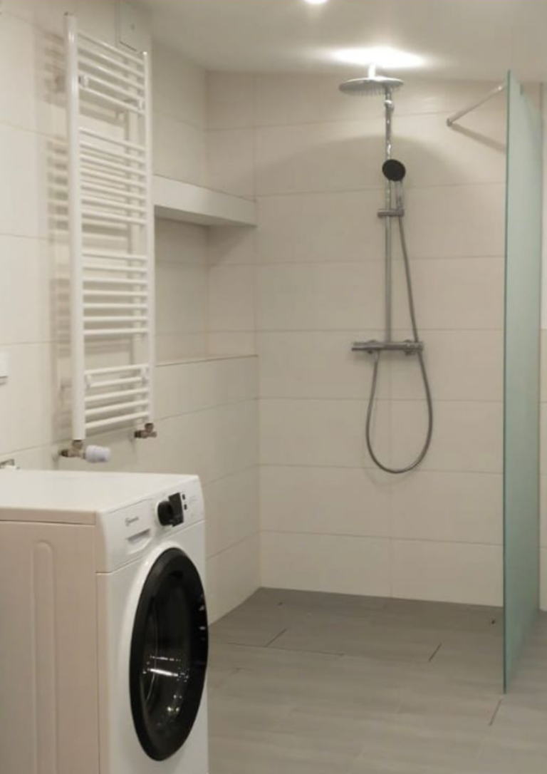 Modernes Badezimmer mit bodengleicher Dusche, Glasabtrennung und Regendusche. Helle Wand- und Bodenfliesen sorgen für ein freundliches Ambiente. Links befindet sich ein weißer Handtuchheizkörper sowie eine Waschmaschine unterhalb einer Ablage-Nische in der Wand.