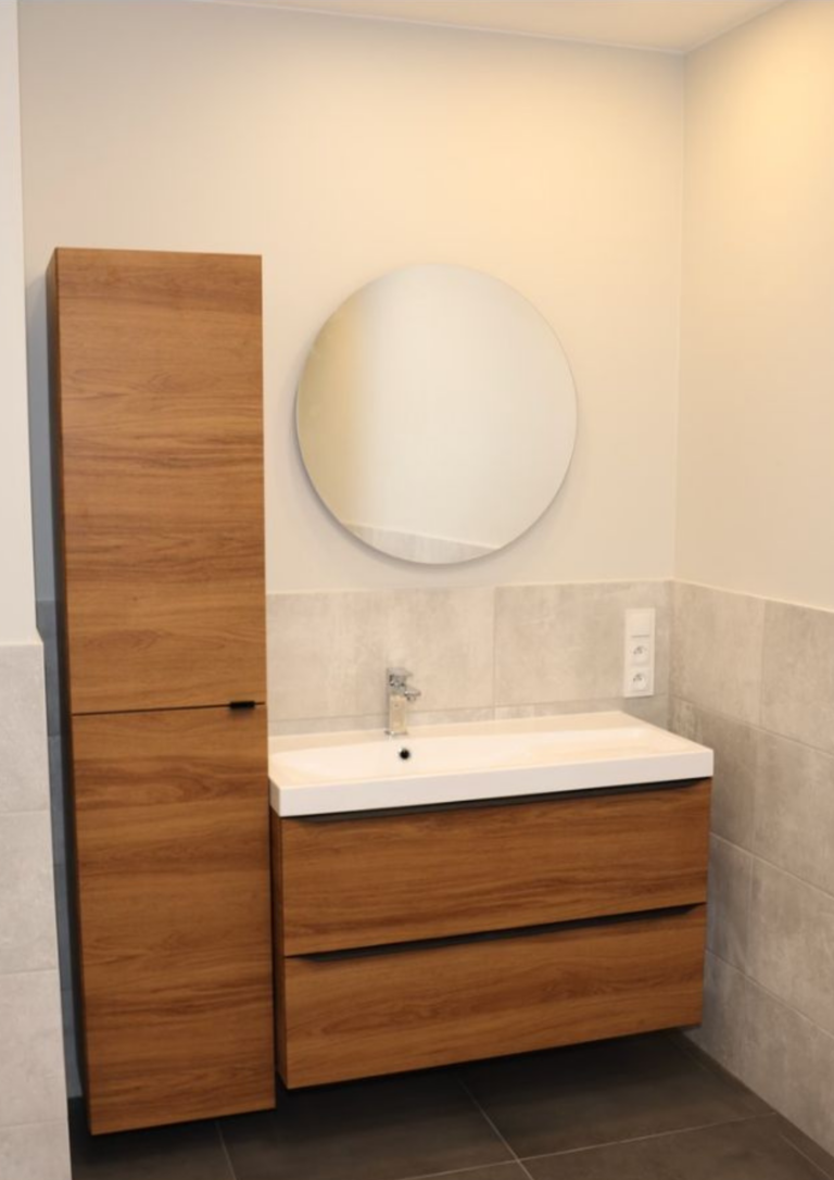 Modernes Badezimmer mit wandhängendem Waschtischunterschrank in warmer Holzoptik, weißem Waschbecken und rundem Spiegel. Links befindet sich ein hoher, schmaler Hochschrank im gleichen Holzdesign, kombiniert mit hellgrauen Wandfliesen und dunklen Bodenfliesen.