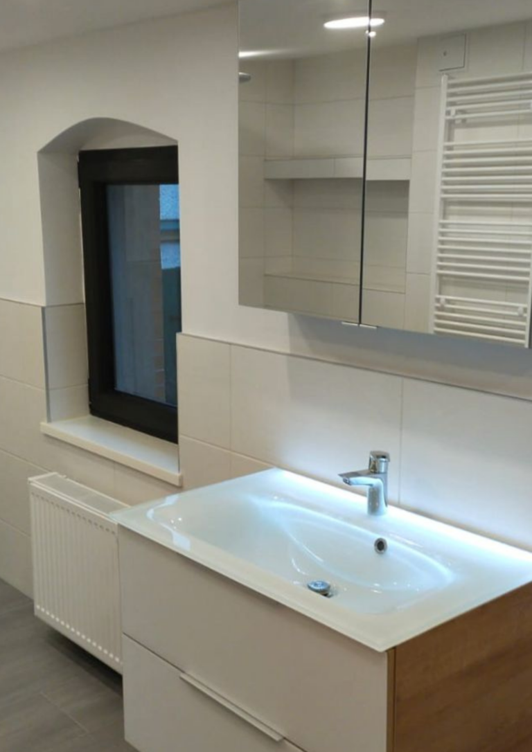 Helles, modern gestaltetes Badezimmer mit weißem Waschtisch und integriertem Unterschrank in Weiß und Holzoptik. Darüber befindet sich ein Spiegelschrank mit Stauraum. Links ein Fenster mit schwarzem Rahmen und darunter ein Heizkörper. Die Wände sind hell gefliest, der Boden in Grau gehalten.