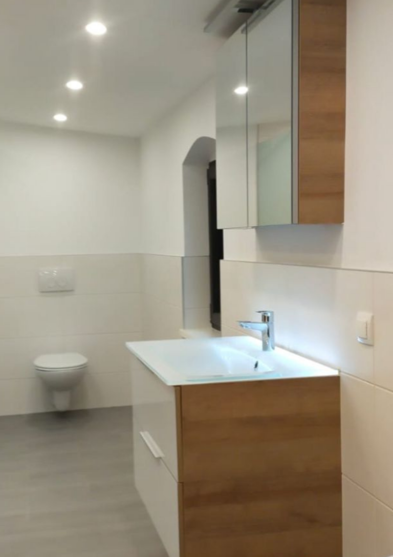 Modernes, helles Badezimmer mit wandhängendem WC, grauem Boden, weiß gefliesten Wänden sowie einem schwebenden Waschbecken-Unterschrank in Holzoptik und weißer Front. Darüber befindet sich ein Spiegelschrank mit integrierter Beleuchtung und Deckenspots sorgen für gleichmäßiges Licht.