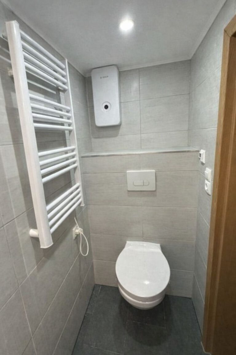 Schmales, modern gefliestes WC mit hellgrauen Wandfliesen und dunklem Boden. Wandhängende Toilette mit weißer Betätigungsplatte, darüber ein elektrischer Durchlauferhitzer. Links befindet sich ein weißer Handtuchheizkörper, rechts eine Holztür. Deckenleuchte sorgt für helles, gleichmäßiges Licht.