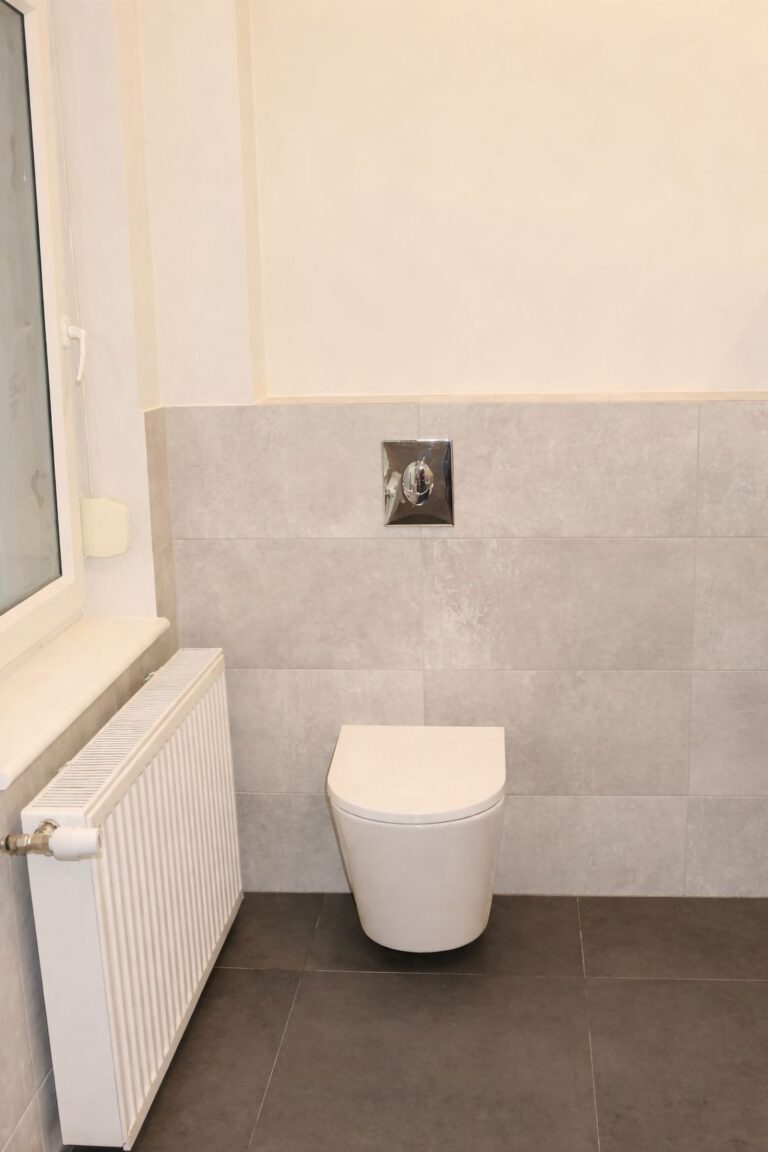 Modernes Gäste-WC mit wandhängender Toilette, grauen Wand- und Bodenfliesen sowie weiß gestrichener Oberwand. Links befindet sich ein Fenster mit Heizkörper darunter, rechts eine WC-Bürste in dekorativem Halter. Die Betätigungsplatte ist in Chrom ausgeführt.